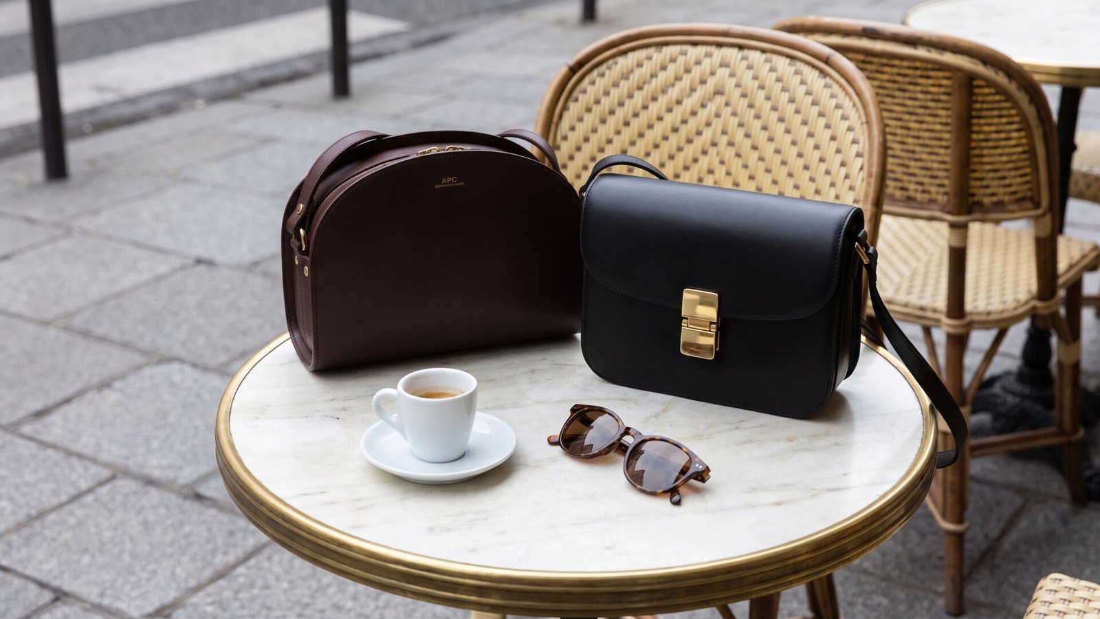 Two minimalist leather crossbody bags, an espresso cup, and sunglasses on a round Parisian café table