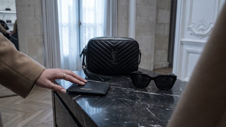 Black Saint Laurent quilted camera bag, leather cardholder, and sunglasses on a dark marble counter
