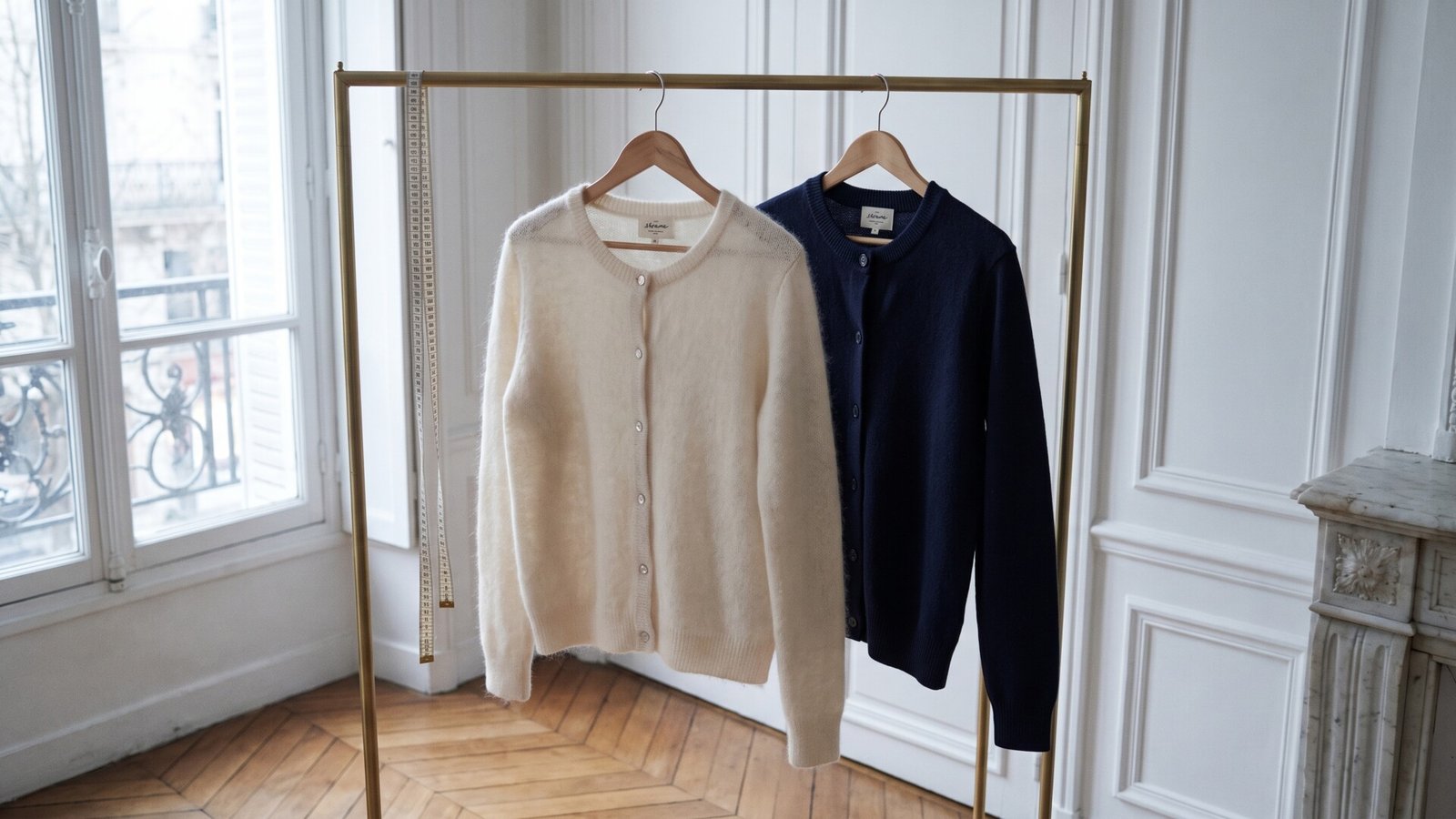 Sézane cream and navy knit cardigans hanging side by side on a brass rack next to a measuring tape